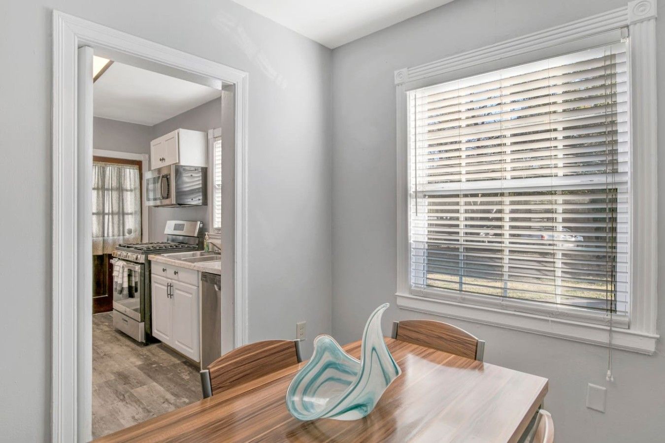 A modern kitchen with a dining area, featuring a large window with blinds, a wooden dining table, and a kitchen island.