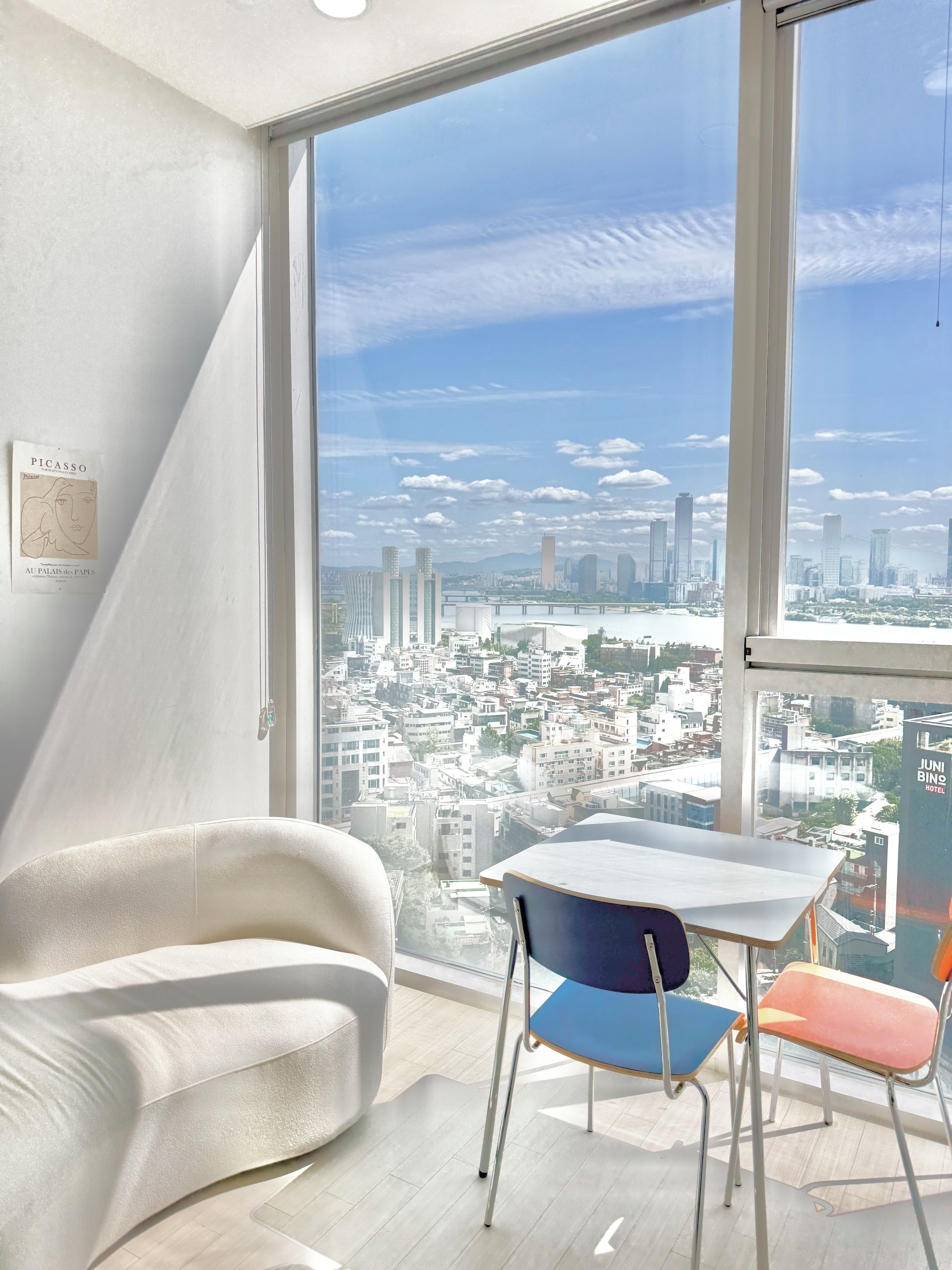A modern hotel room with a large window offering a city view, a white chair, a table, and a framed artwork on the wall.