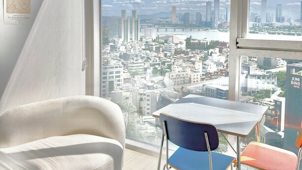 Habitación de hotel moderna con un amplio ventanal que ofrece vistas a la ciudad, una silla blanca, una mesa y una obra de arte enmarcada en la pared.