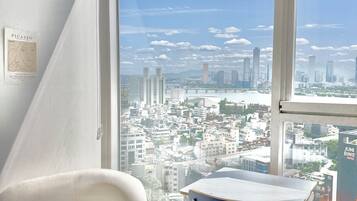A modern hotel room with a large window offering a city view, a white chair, a table, and a framed artwork on the wall.