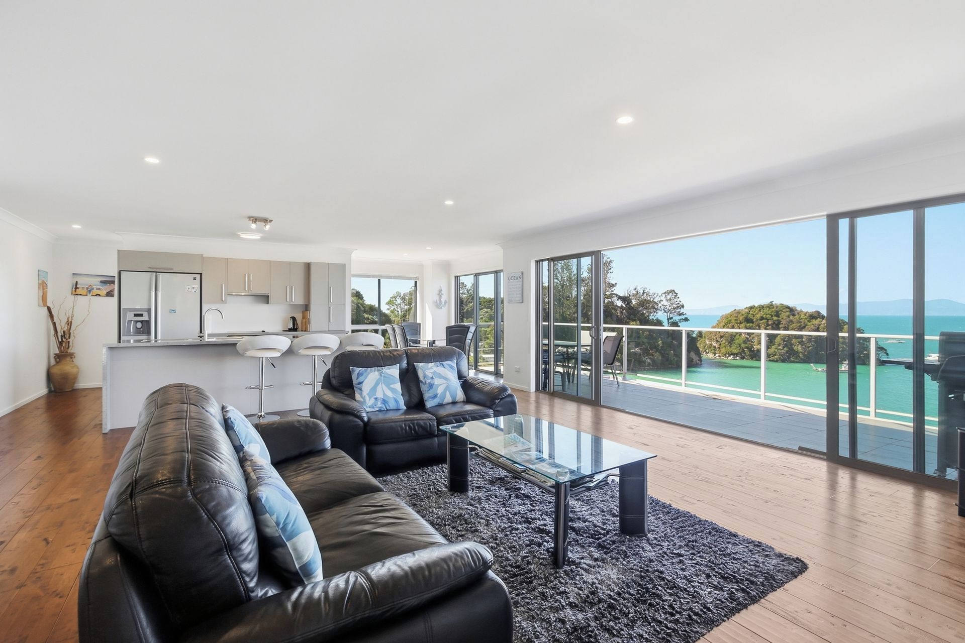 A modern living room with a glass coffee table, a black leather sofa, and a view of the ocean through large sliding doors.