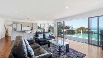 A modern living room with a glass coffee table, a black leather sofa, and a view of the ocean through large sliding doors.