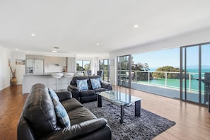 A modern living room with a glass coffee table, a black leather sofa, and a view of the ocean through large sliding doors.