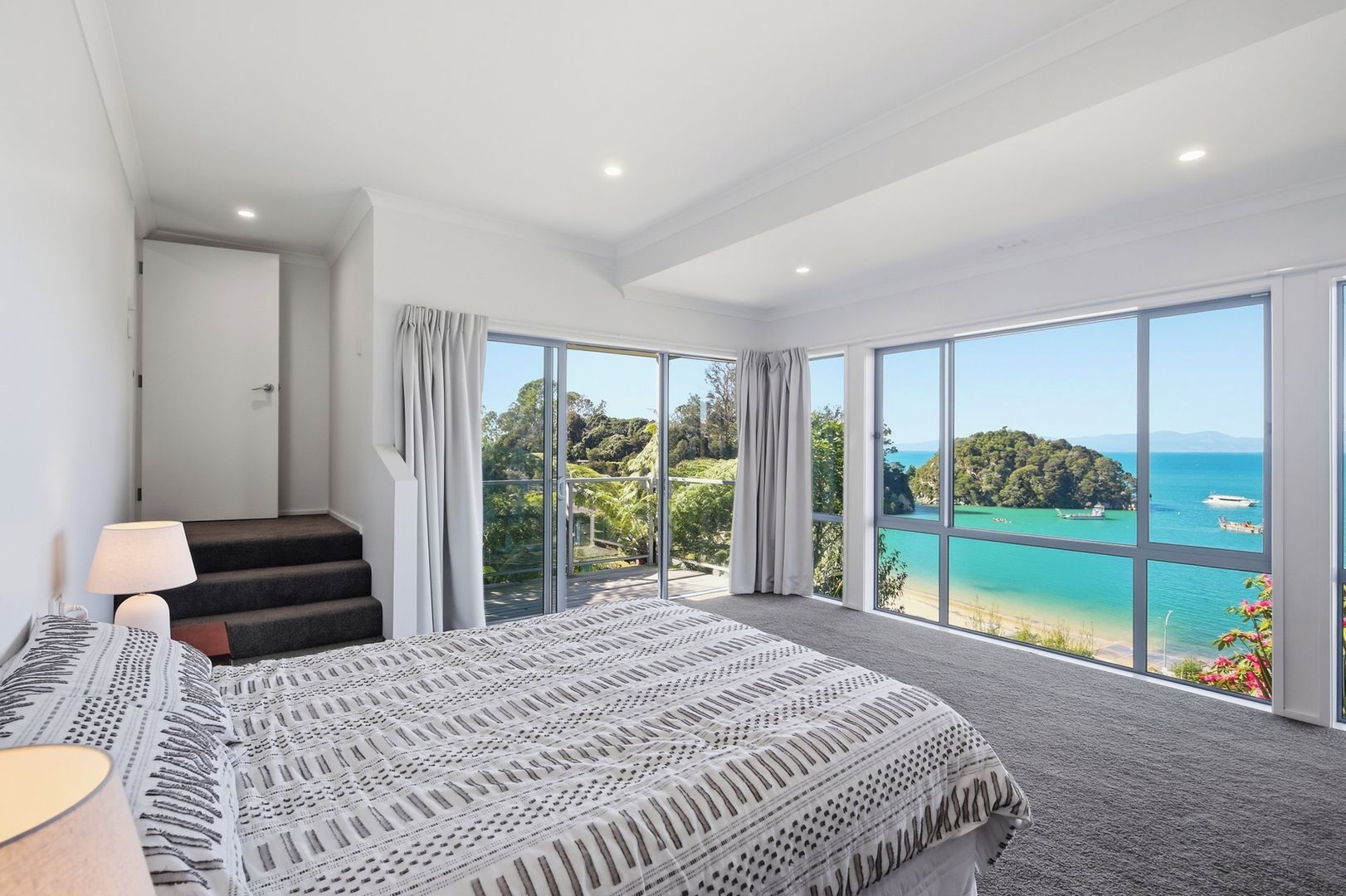 A bedroom with a large bed, a nightstand, a lamp, and a view of a tropical beach and ocean through floor-to-ceiling windows.