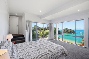 A bedroom with a large bed, a nightstand, a lamp, and a view of a tropical beach and ocean through floor-to-ceiling windows.