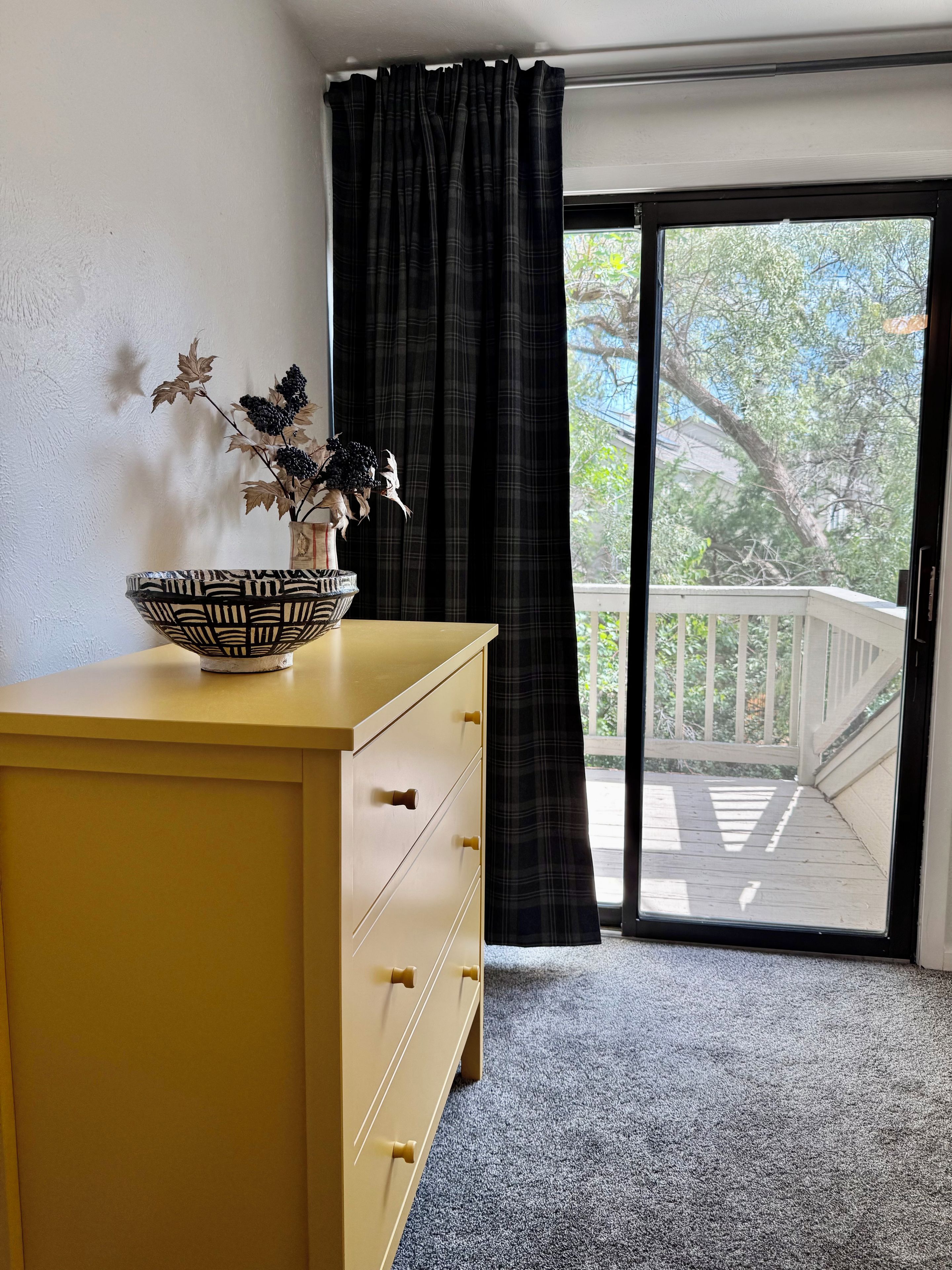 Une pièce avec une commode jaune, un bol décoré posé sur le dessus, une plante en pot et une vue sur un balcon avec une balustrade.