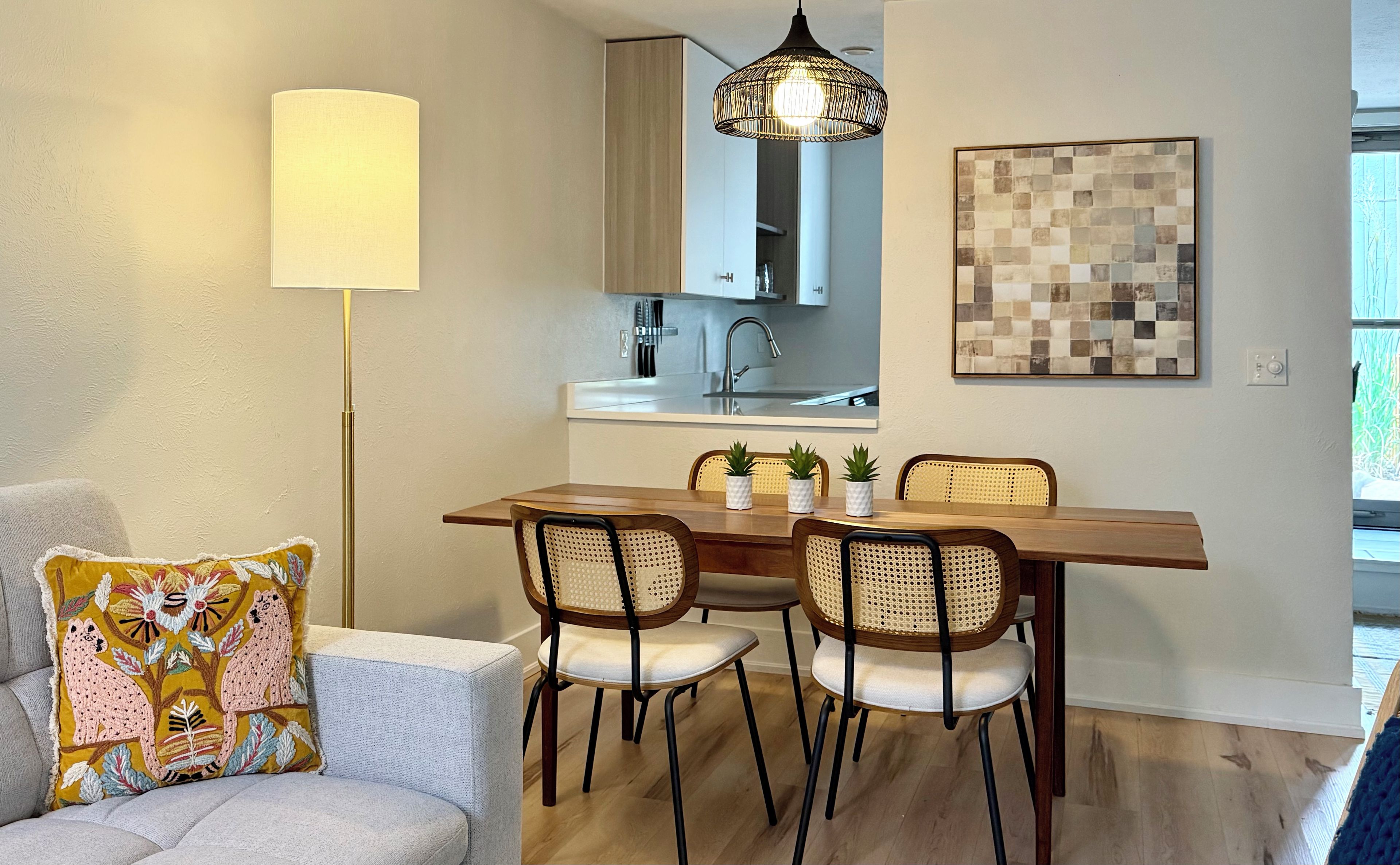 A dining area with a wooden table, four chairs, a pendant light, a kitchen in the background, and a decorative pillow on a couch.