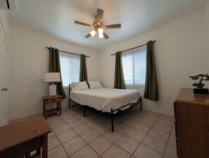 A bedroom with a bed, a nightstand, a lamp, a ceiling fan, and a window with curtains. - Charming 2BR Tucson Home – Minutes to U of A & Downtown! (Tucson)