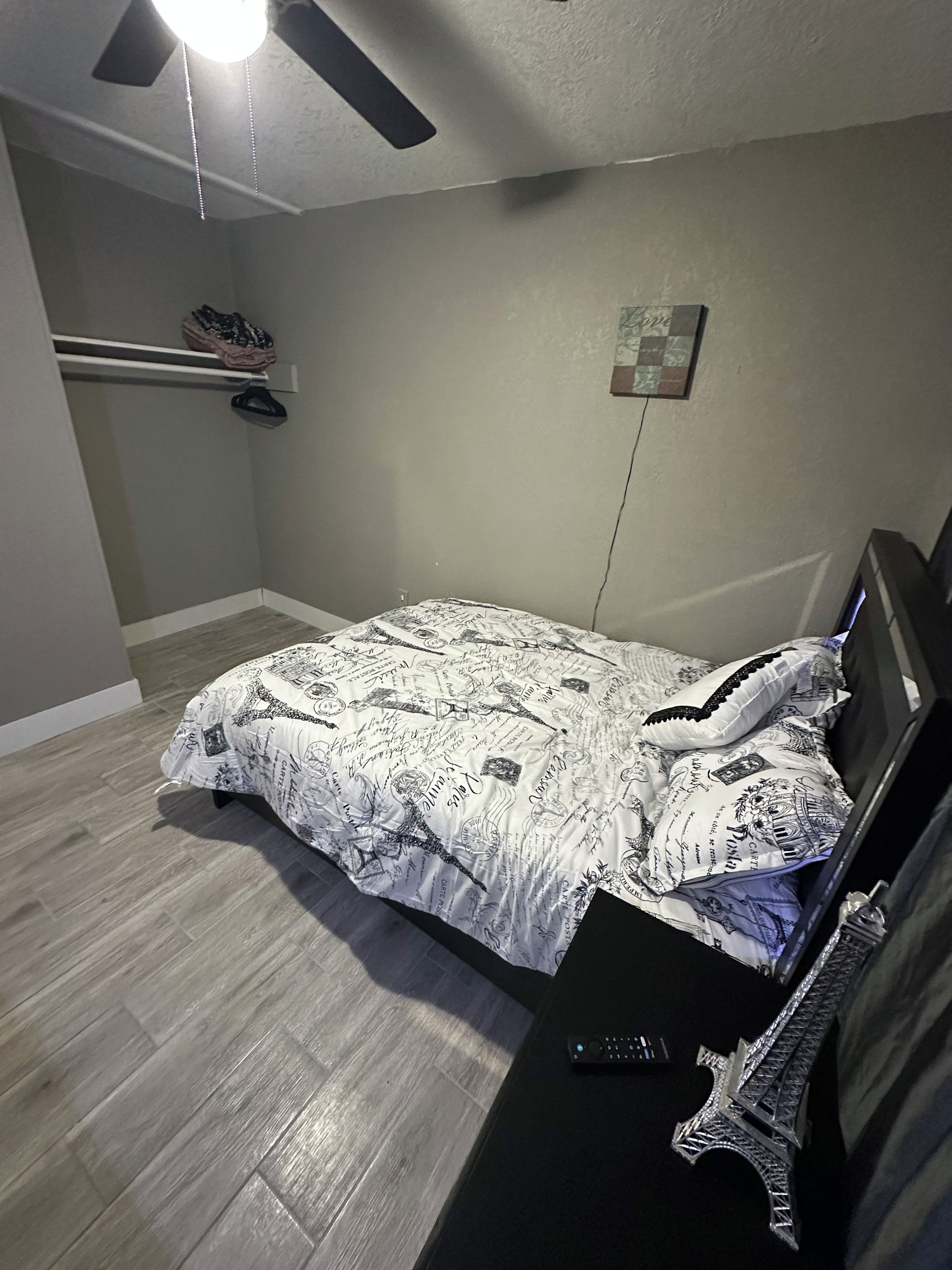 Un dormitori amb un llit, un ventilador de sostre, una prestatgeria penjada a la paret amb sabates i una fotografia petita.