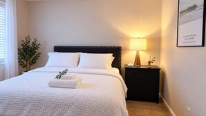 Cama bem arrumada com roupa branca, abajur e um quadro na parede.
