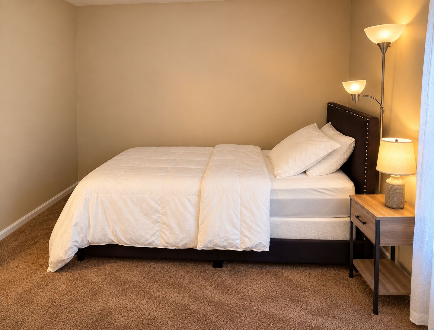 Cama bem arrumada com cabeceira, mesa de cabeceira com luminária e piso coberto por carpete.
