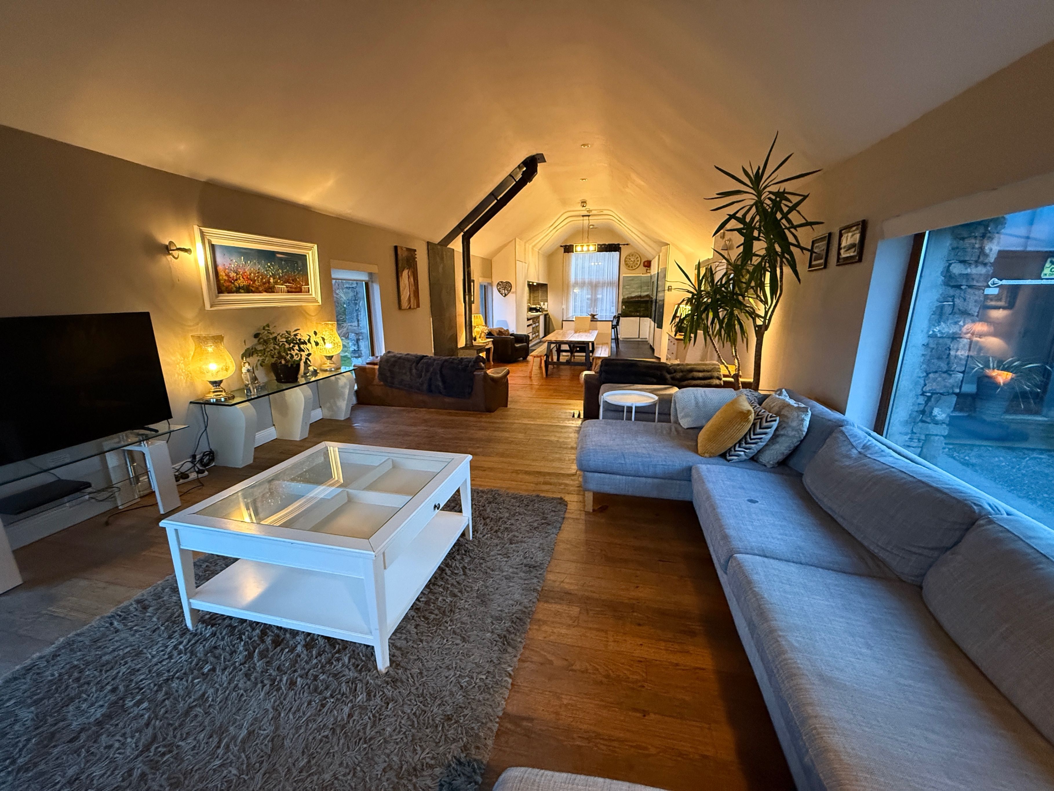 Una sala de estar amplia con un televisor de pantalla plana grande, una mesa de centro de vidrio y un sofá. En el fondo, hay un comedor con una mesa y sillas.