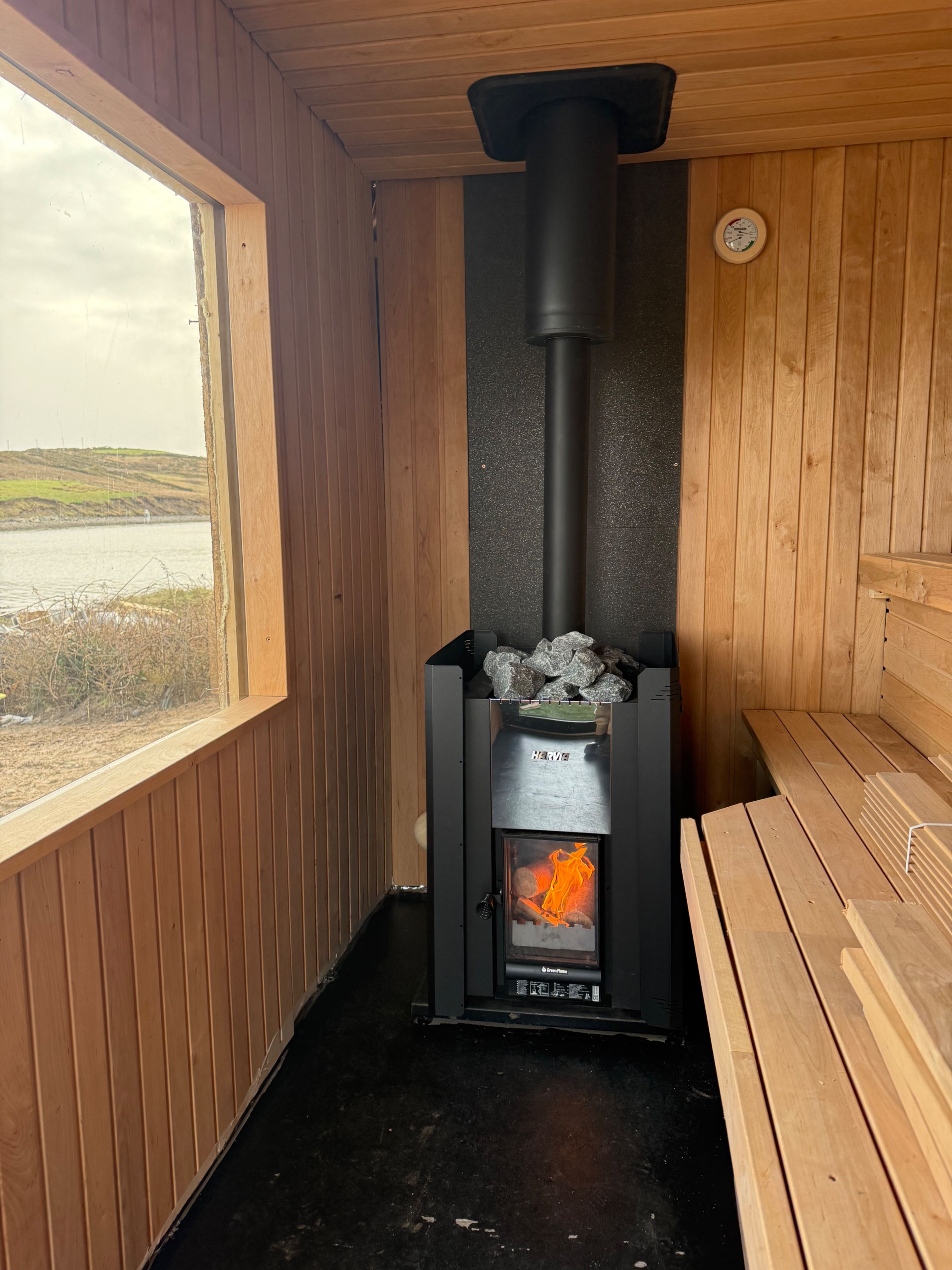 Una sauna con calentador negro, bancos de madera y vista a un lago a través de una ventana.