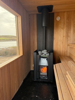 A sauna with a black heater, wooden benches, and a view of a lake through a window. - Private Island Lodge Sleeps 22 with Hot Tub (Westport)