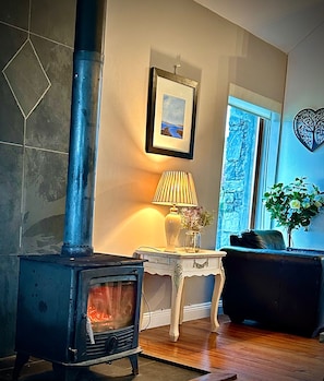 A room with a wood-burning stove, a framed picture on the wall, a lamp on a small table, and a window with a view.