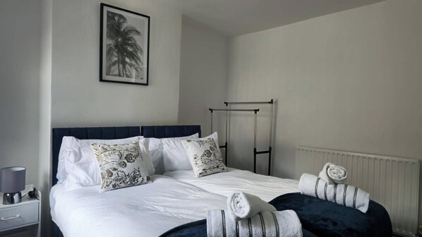 Cama bem arrumada com lençóis brancos e almofadas decorativas, mesa de cabeceira com abajur e um quadro na parede.