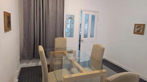 Uma área de refeições com mesa de vidro e quatro cadeiras, cortina cinza e porta com moldura azul.