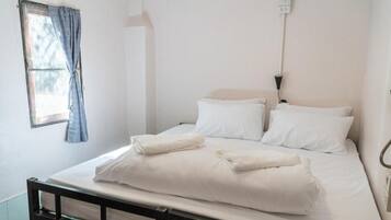 Un lit simple avec du linge de lit blanc, une tête de lit en bois, une fenêtre avec des rideaux bleus et un luminaire fixé au mur.