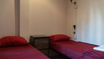A small room with two single beds, each with a red bedspread and matching pillows. There is a small wooden dresser with drawers and a lamp on the wall.