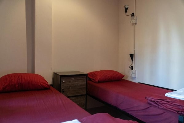 Una habitación pequeña con dos camas individuales, cada una con una colcha roja y almohadas a juego. Hay un pequeño aparador de madera con cajones y una lámpara en la pared.