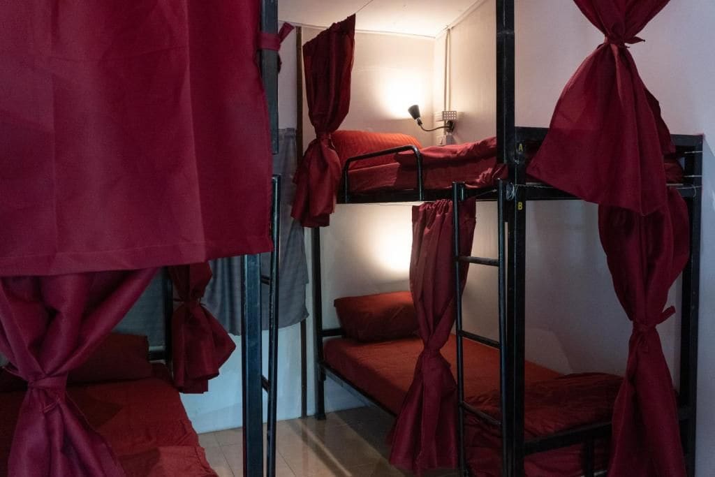 A dormitory room with bunk beds, each with a red bedspread and matching curtains tied at the corners.