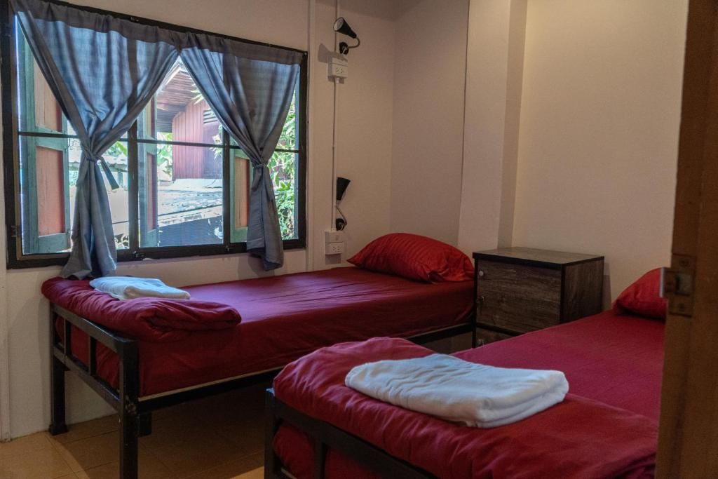 Ein Zimmer mit zwei Einzelbetten, jeweils mit roter Matratze und Kissen, einem hölzernen Nachttisch, einer kleinen Holzkabine und einem Fenster mit Vorhängen.