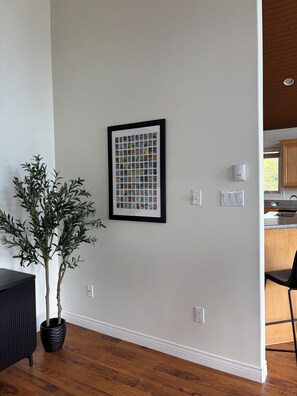 A room with a framed picture on the wall, a potted plant, a wooden floor, and a glimpse of a kitchen area. - Mae’s place l Horse Chops Pond Retreat (Cape Broyle)