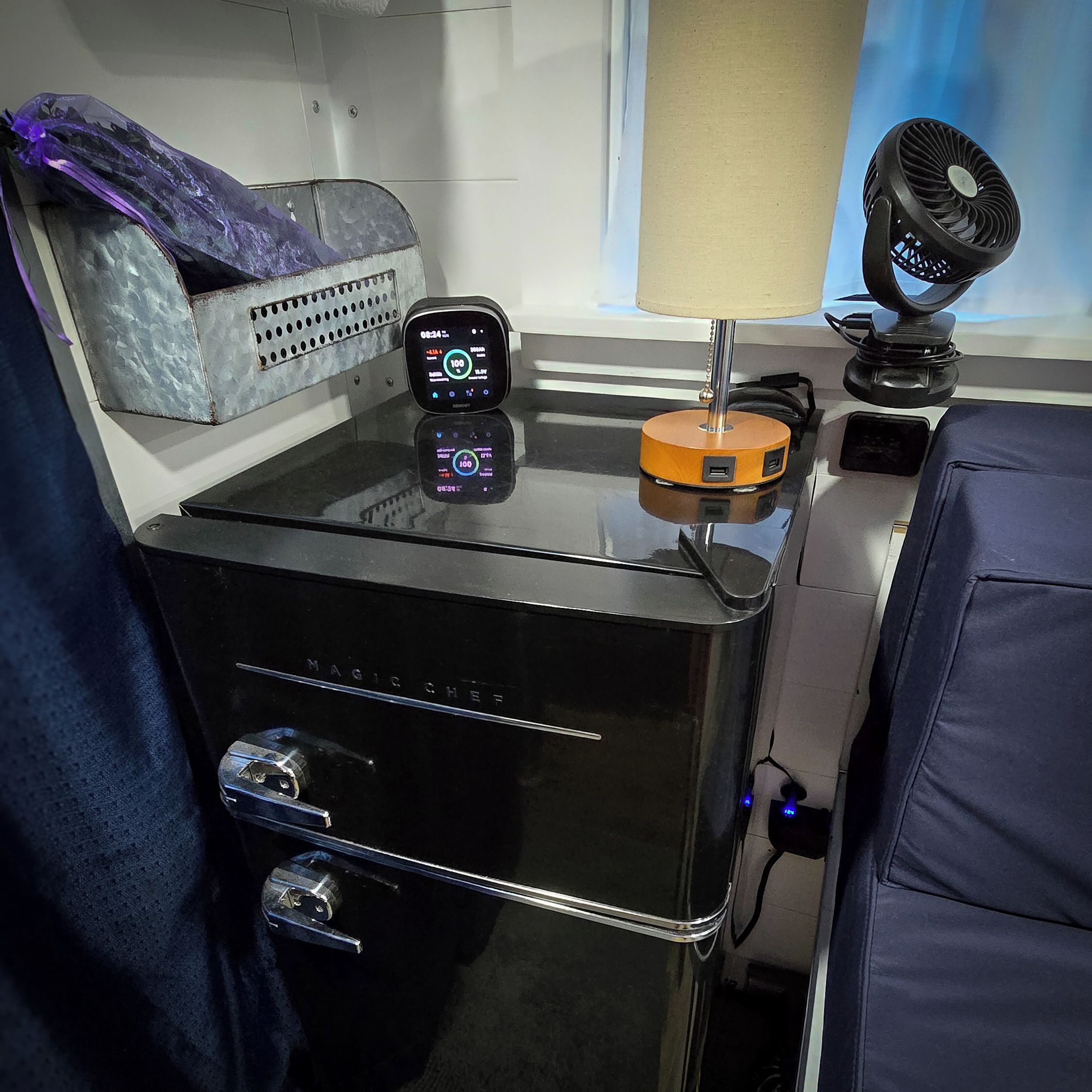 A compact, modern room with a bed, a small table with a lamp, a charging station, and a digital clock.