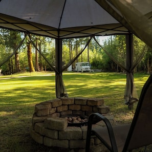 A covered outdoor area with a brick fire pit, a chair, and a camping van in the background.