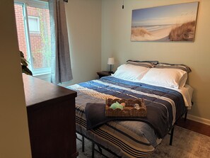 Ein Schlafzimmer mit einem Bett, einem Nachttisch, einer Lampe, einem Fenster mit Vorhängen und einem Gemälde an der Wand.