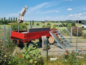 En röd släpvagn med ett överbyggt utrymme, omgiven av ett stängsel av metall, placerad i ett lantligt landskap med åkrar och träd.
