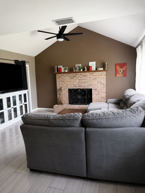 A living room with a brick fireplace, a ceiling fan, and a grey sectional sofa.