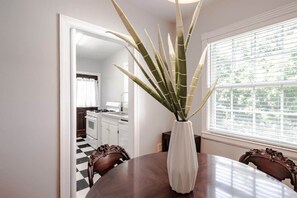 A dining area with a round table, chairs, a white vase with a plant, and a window with blinds. - Sunny Summerville Court Condos (Mobile)