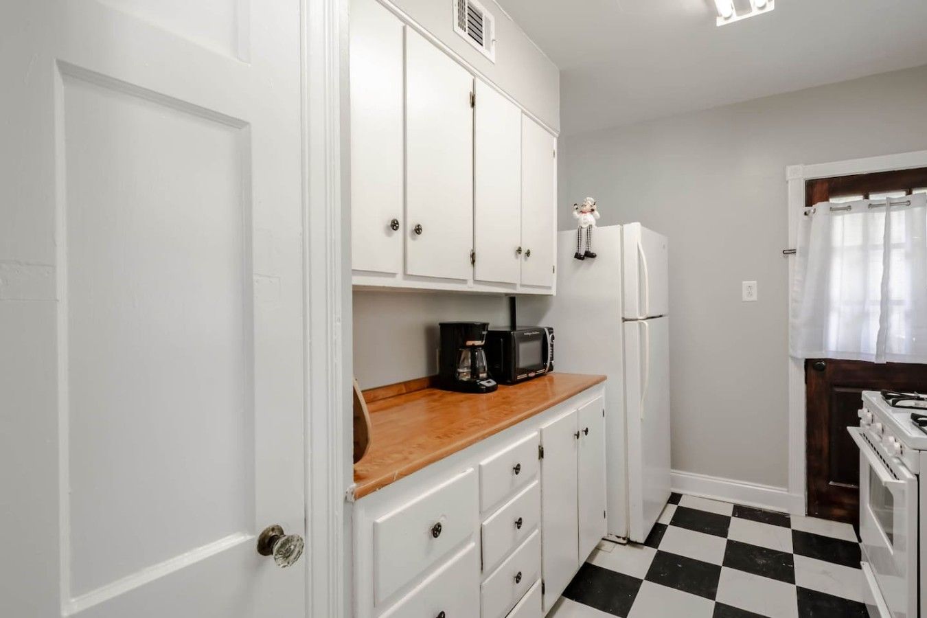 Una cocina compacta con gabinetes blancos, encimera de madera, cafetera negra, refrigerador blanco y piso a cuadros.
