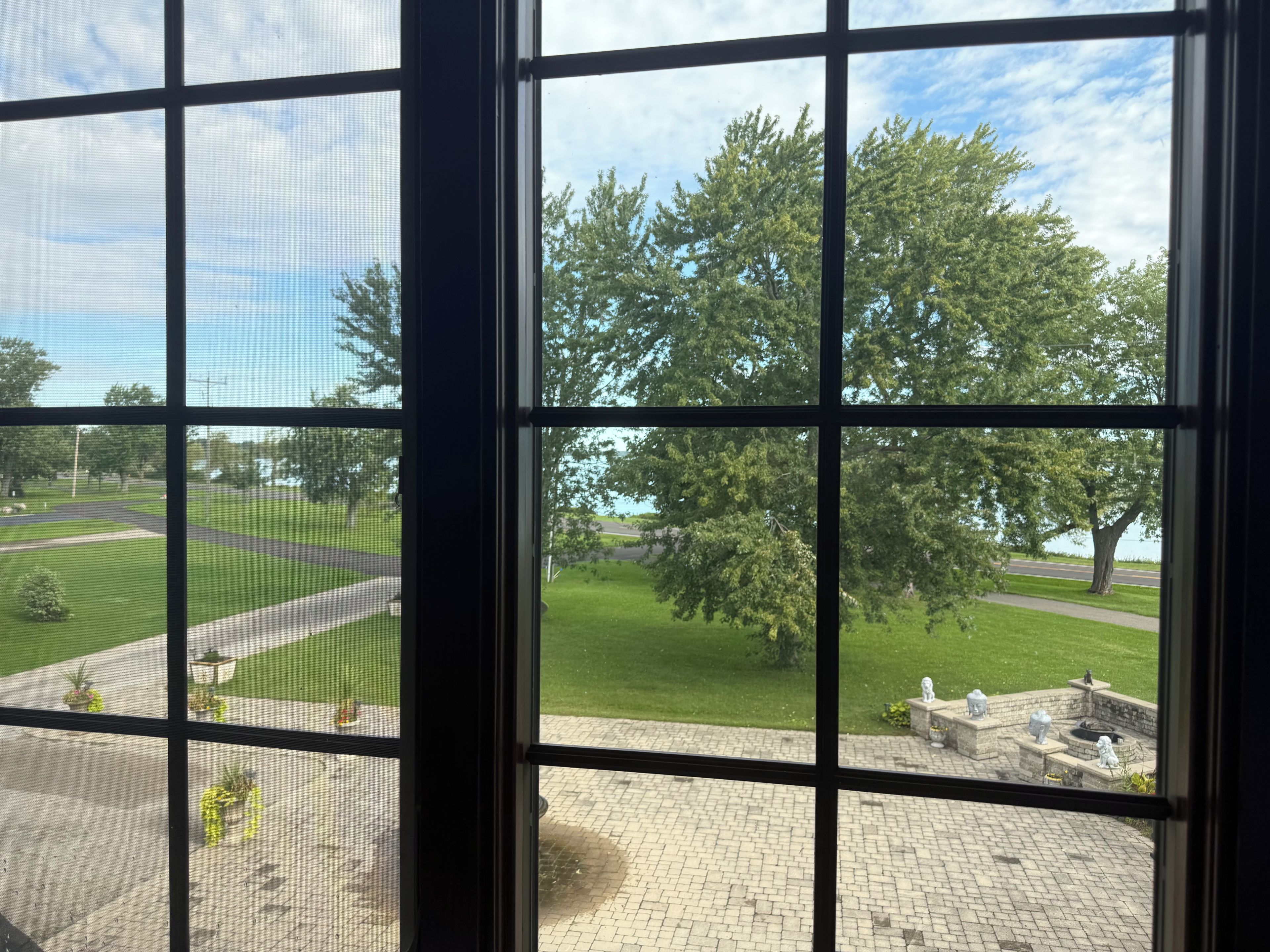 A view through a window with a grid pattern, a paved area with planters, and a grassy field with trees.