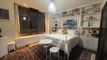 A bedroom with a bed, a window, a radiator, a white chair, a table, a shelf with decorative items, and a painting.