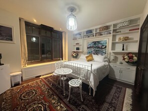 Ein Schlafzimmer mit einem Bett, einem Fenster, einer Heizung, einem weißen Stuhl, einem Tisch, einem Regal mit Dekoration und einem Gemälde.