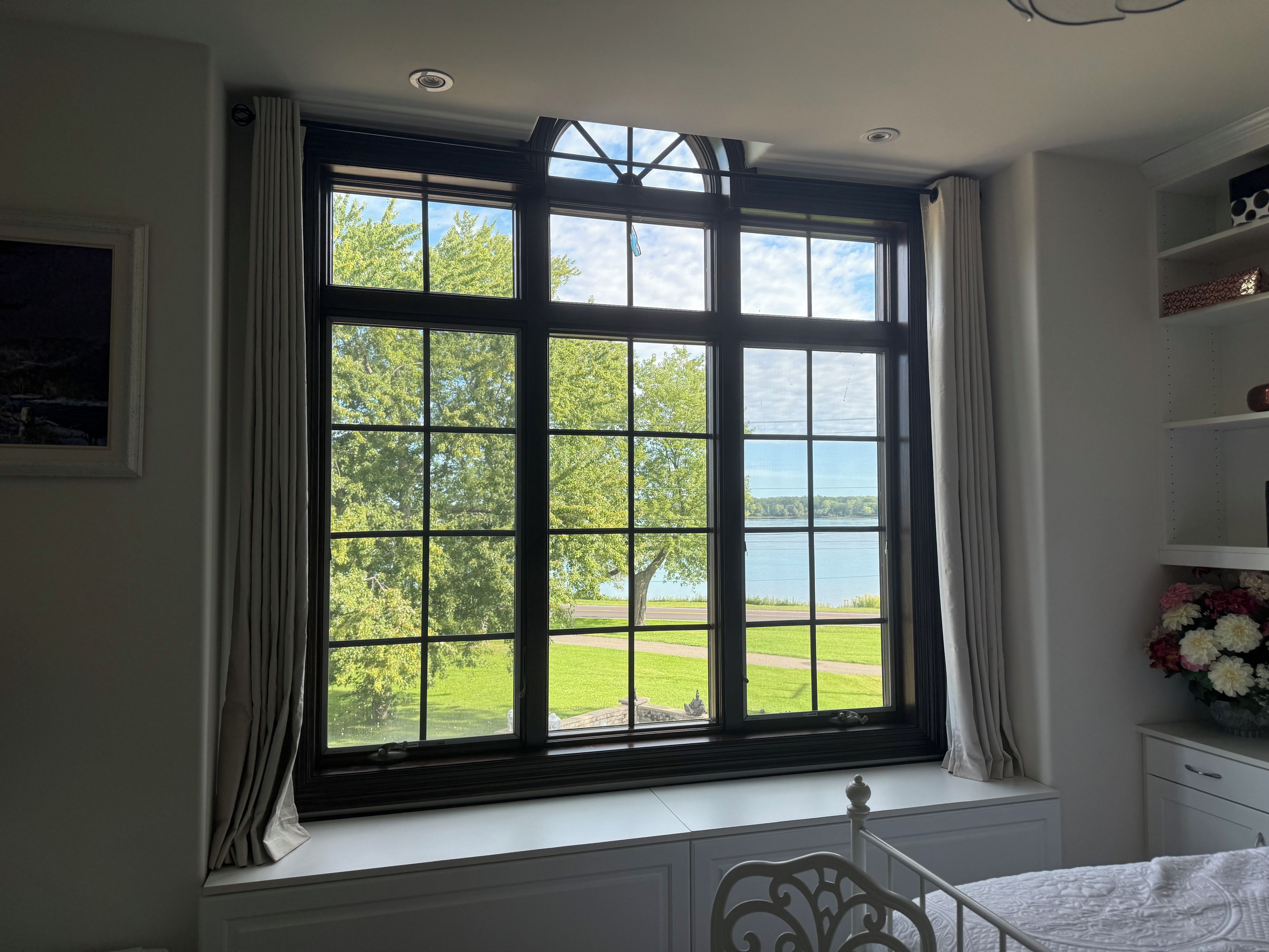 A room with a large window, a bed, a white dresser, and a vase of flowers.