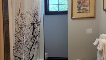 Un baño con cortina de ducha decorada con un diseño de árbol, una ventana con vistas, un cuadro enmarcado en la pared y una alfombra estampada.