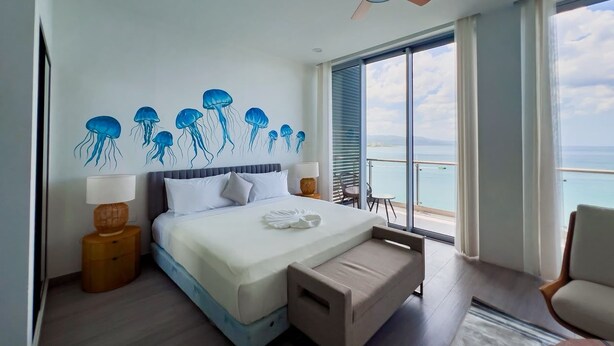 A bedroom with a bed, a ceiling fan, a window with a view of the sea, and a balcony with a chair. - The Bloom - Oceanfront King Suite (Montego Bay)