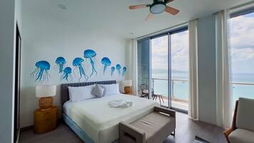 A bedroom with a bed, a ceiling fan, a window with a view of the sea, and a balcony with a chair.
