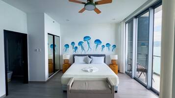 Une chambre moderne dotée d’un grand lit, d’un ventilateur de plafond et offrant une vue sur la mer grâce à de grandes fenêtres du sol au plafond.
