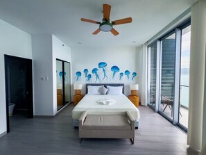 Un dormitori modern amb un llit gran, un ventilador de sostre i vistes al mar a través de grans finestres de sostre a terra.