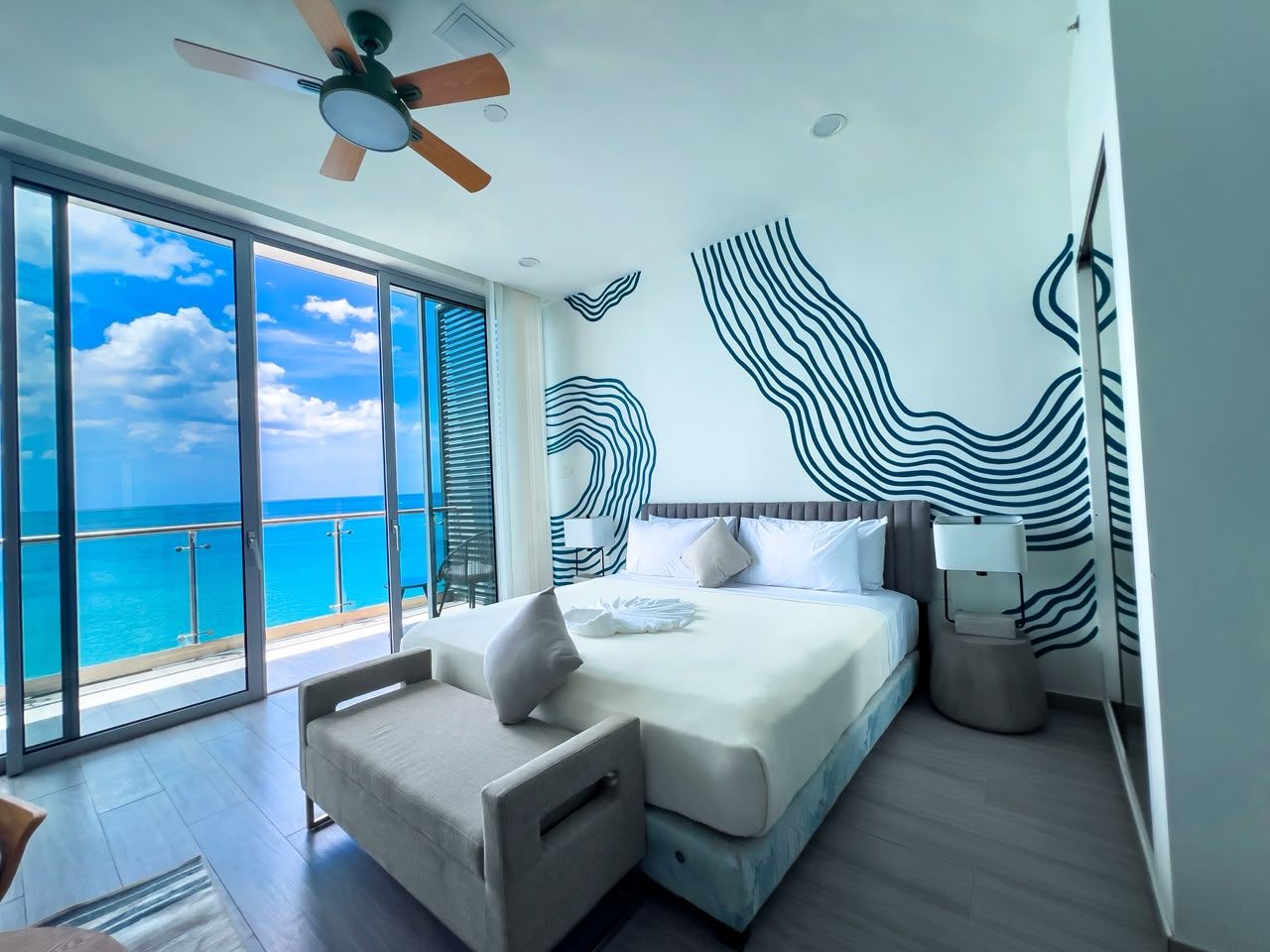 A modern hotel room with a large bed, a seating area, a ceiling fan, and a view of the ocean.