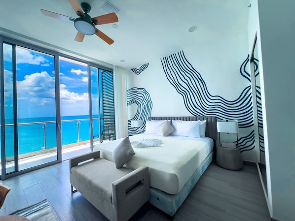 Una habitación de hotel moderna con una cama grande, zona de estar, ventilador de techo y vista al océano.