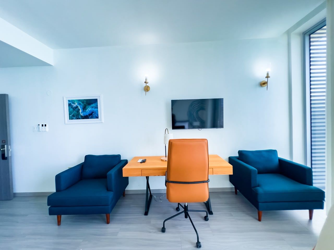 Een moderne kamer met een bureau, twee blauwe banken, een oranje bureaustoel, een flatscreen-tv en wandlampen.