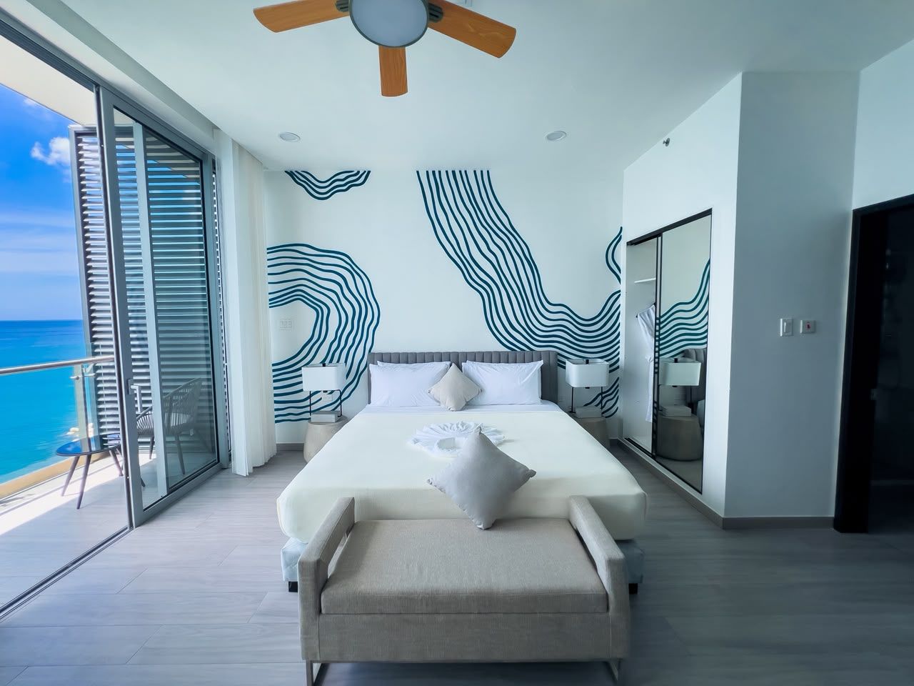 A modern bedroom with a large bed, a ceiling fan, and a view of the ocean.