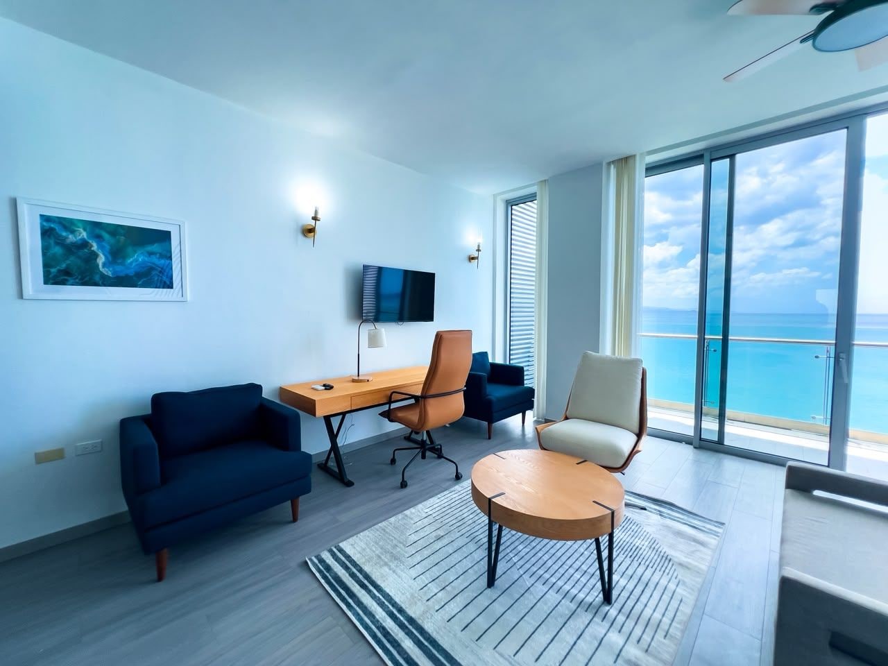 A modern living room with a large window, a flat-screen TV, a desk, and seating arrangements.