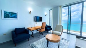A modern living room with a large window, a flat-screen TV, a desk, and seating arrangements. - The Tides - Oceanfront King Suite (Montego Bay)