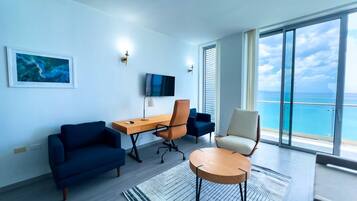 A modern living room with a large window, a flat-screen TV, a desk, and seating arrangements.